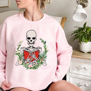 Pode incluir: Camisola rosa claro com um esqueleto lendo um livro vermelho, rodeado por uma coroa de folhas verdes e flores. Os bra&ccedil;os do esqueleto est&atilde;o cruzados sobre o livro. A camisola &eacute; de gola redonda.