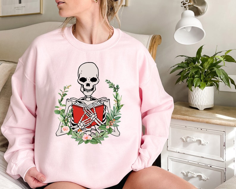Pode incluir: Camisola rosa claro com um esqueleto lendo um livro vermelho, rodeado por uma coroa de folhas verdes e flores. Os bra&ccedil;os do esqueleto est&atilde;o cruzados sobre o livro. A camisola &eacute; de gola redonda.