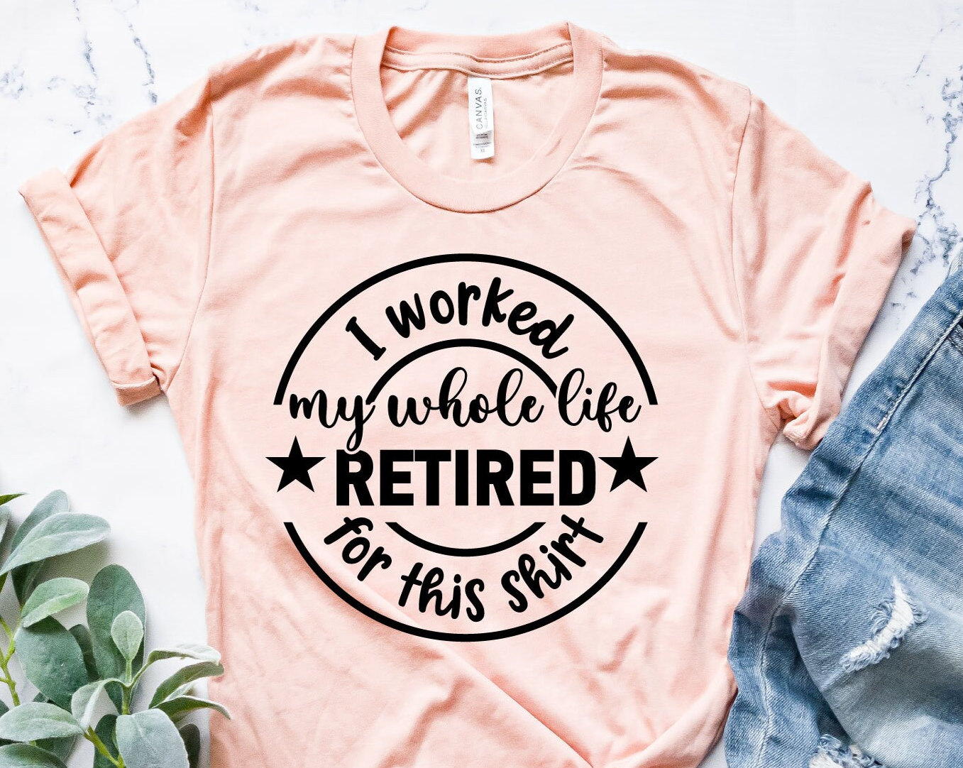 Pode incluir: Uma t-shirt rosa claro com o texto preto "I worked my whole life RETIRED for this shirt" num design circular. A t-shirt tem mangas enroladas e é feita de um material macio. Combinada com calças de ganga azuis.