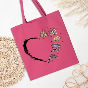 May include: A pink tote bag featuring a black heart design with illustrated books. The books are in various colors and styles, with some open and others stacked. The bag has long handles for carrying.