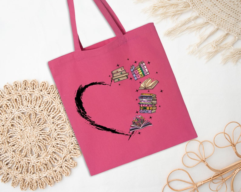 May include: A pink tote bag featuring a black heart design with illustrated books. The books are in various colors and styles, with some open and others stacked. The bag has long handles for carrying.