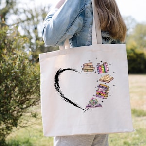 May include: Cream-colored tote bag featuring a black heart shape with a brushstroke design. Inside the heart are colorful illustrations of stacked books, open books, and stars. The bag has long straps.