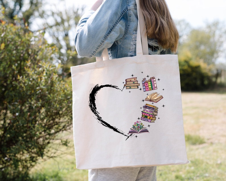 May include: Cream-colored tote bag featuring a black heart shape with a brushstroke design. Inside the heart are colorful illustrations of stacked books, open books, and stars. The bag has long straps.