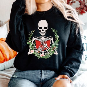 Pode incluir: Camisola preta com um esqueleto lendo um livro vermelho, rodeado por uma coroa de flores. O esqueleto tem um cr&acirc;nio branco e segura o livro com as m&atilde;os &oacute;sseas. O design combina elementos assustadores e florais.