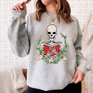 Pode incluir: Um moletom cinza claro com um esqueleto lendo um livro vermelho, cercado por uma coroa de folhas verdes e flores rosa. O design do esqueleto &eacute; em branco e preto. O moletom tem mangas compridas.