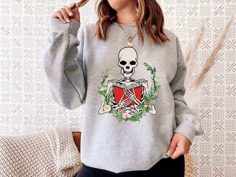 Pode incluir: Um moletom cinza claro com um esqueleto lendo um livro vermelho, cercado por uma coroa de folhas verdes e flores rosa. O design do esqueleto &eacute; em branco e preto. O moletom tem mangas compridas.