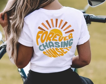May include: White t-shirt with a circular graphic on the back featuring a yellow sun and the words "Forever Chasing Sunsets" in orange and teal. The design includes brown and orange sun rays.