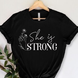 May include: Black t-shirt with the white text "She is STRONG". The design includes a floral illustration with a hand holding the flowers. The shirt is hanging on a wooden hanger.