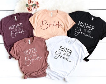 May include: Five short-sleeved t-shirts in various colors: burgundy, peach, black, mauve, and white. Each shirt features a different title in a script font: "Mother of the Bride", "Bride", "Mother of the Groom", "Sister of the Bride", and "Sister of the Groom".