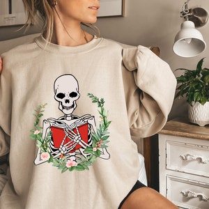 Pode incluir: Camisola bege com um esqueleto lendo um livro vermelho, rodeado por uma coroa de flores. O esqueleto &eacute; em preto e branco, o livro &eacute; vermelho. A coroa inclui folhas verdes e flores cor-de-rosa, criando um contraste marcante.