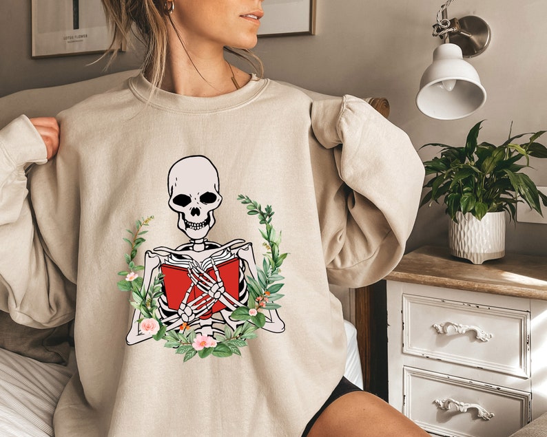 Pode incluir: Camisola bege com um esqueleto lendo um livro vermelho, rodeado por uma coroa de flores. O esqueleto &eacute; em preto e branco, o livro &eacute; vermelho. A coroa inclui folhas verdes e flores cor-de-rosa, criando um contraste marcante.