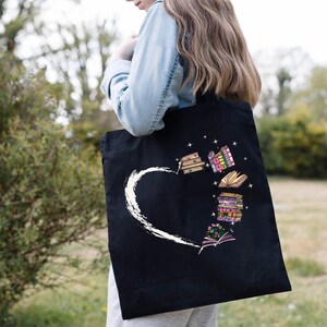 May include: Black tote bag featuring a white heart design with a brushstroke effect. The bag showcases colorful illustrations of books, some open, some stacked, and others with floral accents, creating a literary theme.