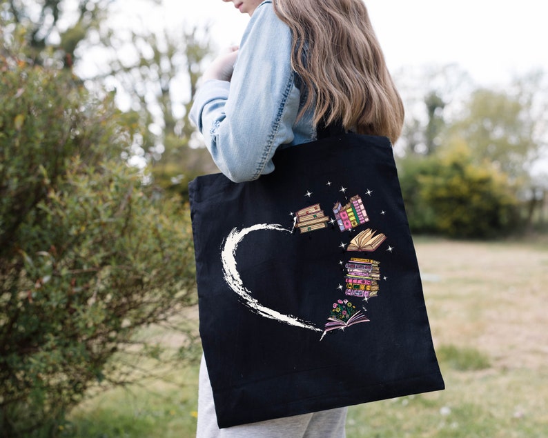 May include: Black tote bag featuring a white heart design with a brushstroke effect. The bag showcases colorful illustrations of books, some open, some stacked, and others with floral accents, creating a literary theme.