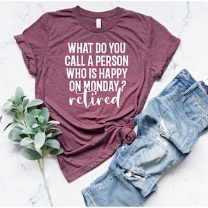Pode incluir: Uma camiseta bordô com o texto "WHAT DO YOU CALL A PERSON WHO IS HAPPY ON MONDAY? retired" em branco. A camiseta é combinada com jeans azuis e um raminho de folhas verdes.
