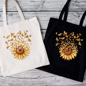 Pode incluir: Duas sacolas, uma na cor branco sujo e outra preta, cada uma com uma estampa gráfica de um grande girassol amarelo com inúmeras pequenas borboletas amarelas que parecem emergir do centro e voar para cima em um padrão disperso.