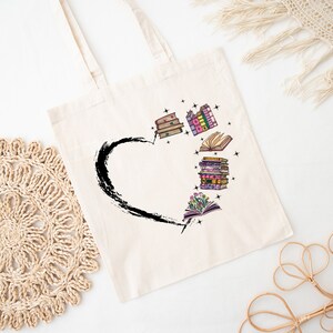 May include: Cream-colored tote bag featuring a black heart-shaped brushstroke design. The heart is filled with colorful illustrations of books and flowers. The bag has two handles and is made of a light-colored fabric.