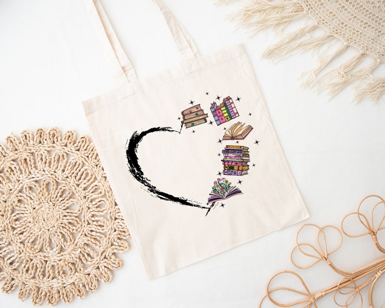 May include: Cream-colored tote bag featuring a black heart-shaped brushstroke design. The heart is filled with colorful illustrations of books and flowers. The bag has two handles and is made of a light-colored fabric.