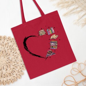 May include: A red tote bag featuring a black heart shape with a stack of books and open books with flowers. The books are in various colors and styles. The tote bag has two red handles.