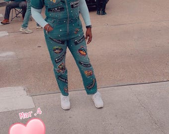 May include: A woman wearing a teal denim jumpsuit with various patches and embellishments. The jumpsuit has a zip fastening and a collared neckline. The woman is standing on a pavement with a heart-shaped graphic in the foreground.