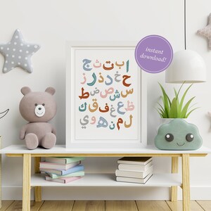 Peut inclure: Une impression encadrée blanche avec des lettres de l'alphabet arabe colorées sur un fond blanc. Les lettres sont disposées en colonne verticale. L'impression est accrochée à un mur blanc avec un ours en peluche, une étoile et un croissant de lune sur une étagère en dessous.