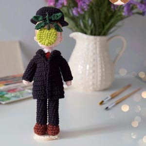 Puede incluir: Muñeca de ganchillo inspirada en el cuadro "El hijo del hombre" de René Magritte. La muñeca presenta un abrigo negro, un bombín y una manzana verde que cubre la cara. La muñeca está frente a un jarrón con flores.