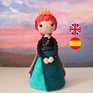 Puede incluir: Una muñeca de crochet que lleva un vestido negro y verde con una capa morada y naranja. La muñeca tiene el pelo rojo y una corona dorada.