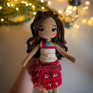 May include: Crocheted doll with brown hair, a white top with a red design, and a red skirt with white accents. The doll is wearing a blue necklace with a turquoise pendant.