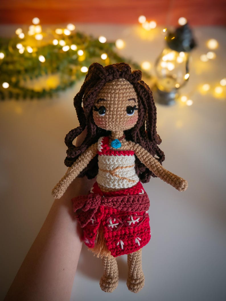 May include: Crocheted doll with brown hair, a white top with a red design, and a red skirt with white accents. The doll is wearing a blue necklace with a turquoise pendant.