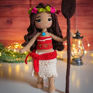 May include: A crocheted doll with brown hair, wearing a red and white top, a white skirt, and a flower crown. The doll is holding a brown wooden spear.