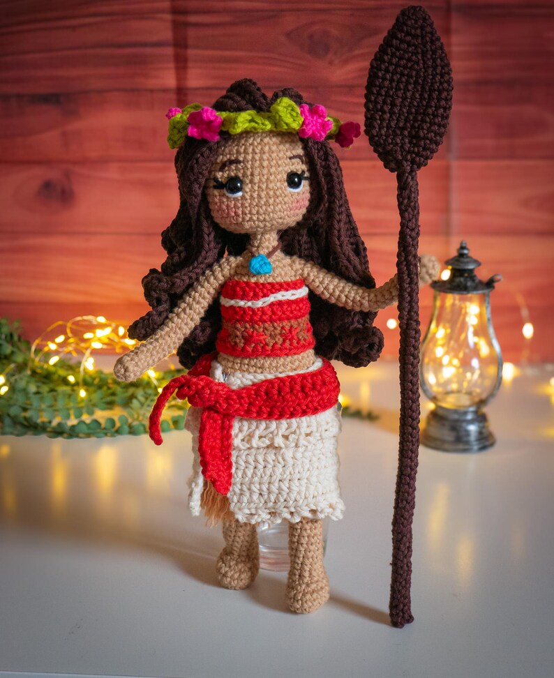 May include: A crocheted doll with brown hair, wearing a red and white top, a white skirt, and a flower crown. The doll is holding a brown wooden spear.