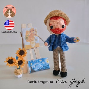 Puede incluir: Muñeco amigurumi de crochet de Vincent Van Gogh, con una chaqueta azul, pantalones marrones y un sombrero marrón. El muñeco sostiene un pincel y una paleta de pinturas. El muñeco está de pie frente a un caballete de madera con dos pinturas, una de girasoles y otra de una noche estrellada.