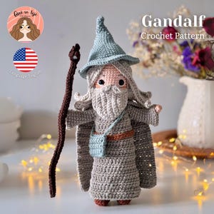 Puede incluir: Muñeco de Gandalf tejido a ganchillo, con una túnica gris, un sombrero azul y un largo bastón marrón. El muñeco tiene una larga barba blanca y sostiene una pequeña bolsa azul. Se muestran las palabras "Gandalf Crochet Pattern".
