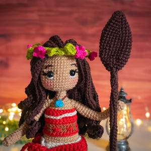 May include: A crocheted doll with brown hair, a flower crown, and a red and white top. The doll is holding a brown crocheted spear.