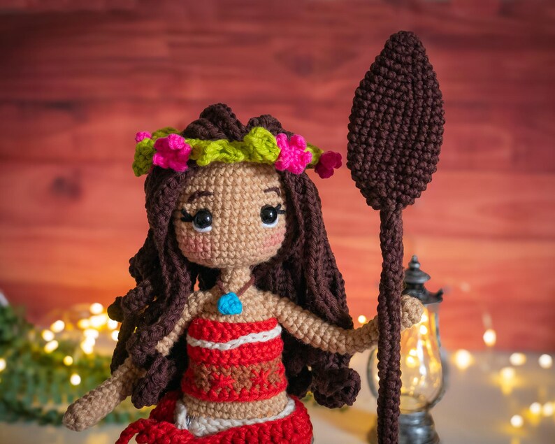 May include: A crocheted doll with brown hair, a flower crown, and a red and white top. The doll is holding a brown crocheted spear.