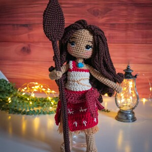 May include: Crocheted doll with brown hair, wearing a red and white dress and holding a brown spear. The doll is standing on a white surface.