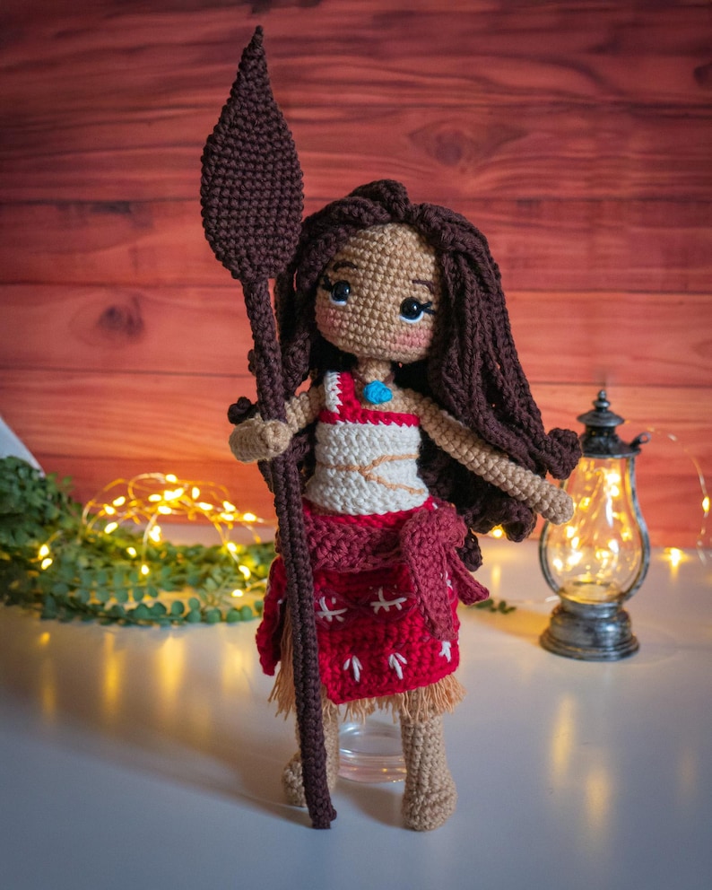 May include: Crocheted doll with brown hair, wearing a red and white dress and holding a brown spear. The doll is standing on a white surface.