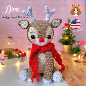 Może przedstawiać: Wzór amigurumi renifera na szydełku o nazwie "Dixie". Renifer jest brązowy i biały z czerwonym szalikiem. Wzór jest dostępny w języku angielskim.