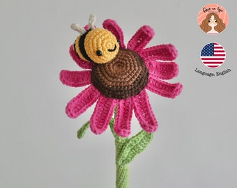 May include: A crocheted pink flower with a brown center and a yellow and black crocheted bee sleeping on top. The flower is on a green stem.
