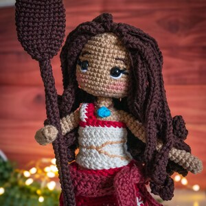 May include: Crocheted doll with brown hair, wearing a white and red dress and holding a brown spear. The doll has a blue jewel on its chest.