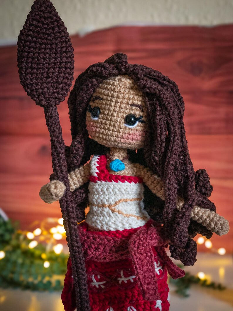 May include: Crocheted doll with brown hair, wearing a white and red dress and holding a brown spear. The doll has a blue jewel on its chest.