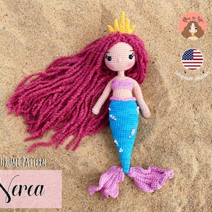 May include: Crocheted amigurumi mermaid doll with pink hair, a blue tail, and a gold crown. The doll is lying on a sandy beach. The doll is named "Nerea" and is part of an amigurumi pattern.