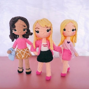 Puede incluir: Tres muñecas de crochet con cabello rubio y castaño que llevan suéteres y faldas rosas. Las muñecas están de pie sobre una superficie rosa.
