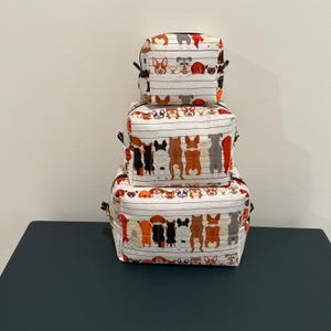 May include: Three stacked, square fabric storage containers with a dog-themed pattern. The containers are white with horizontal stripes and feature various dog breeds in different colors. Each container has a black zipper.