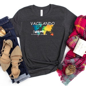May include: Dark gray t-shirt with the words "VACILANDO" and "Welcome to nowhere in particular" printed on the front. The design includes a colorful world map. The shirt is paired with blue jeans and tan sandals.