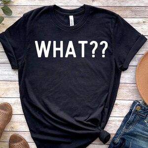 May include: Black t-shirt with white text that says "WHAT??" on a wooden background.