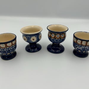 May include: Four ceramic egg cups with a pedestal base, featuring a cream-colored interior and a blue exterior decorated with a repeating pattern of brown and white circular motifs and blue floral accents around the base of each cup.