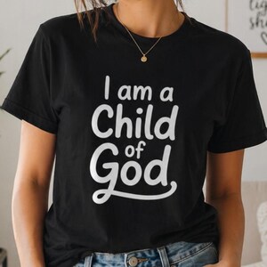 以下が含まれることがあります： 白い文字で「I am a Child of God」と書かれた黒いクルーネックTシャツ。テキストは太字の様式化されたフォントです。シャツは柔らかい素材でできており、カジュアルウェアに適しています。