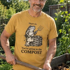 Op de afbeelding: Mosterdgeel T-shirt met een grappige afbeelding van een kikker met een hooivork, naast een compostbak. De tekst op het shirt luidt: "That's it. You're going in the COMPOST."