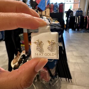 May include: Gold-coloured angel earrings with clear gemstone details. The earrings are presented on a white card, with the text ".14 KT GOLD". The earrings are held by a person in a retail environment.
