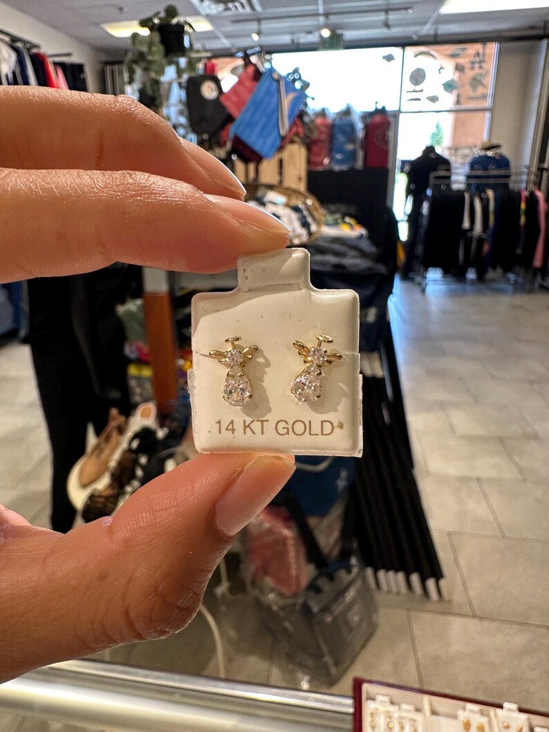May include: Gold-coloured angel earrings with clear gemstone details. The earrings are presented on a white card, with the text ".14 KT GOLD". The earrings are held by a person in a retail environment.